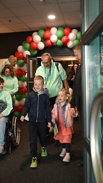Kinderen en vrijwilligers lopen een feestelijke ruimte binnen tijdens een maatschappelijke impactdag, met rood-wit-groene ballonnen en deelnemers in rolstoelen op de achtergrond.